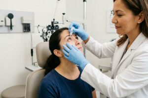 An optometrist in gloves applies eye drops to a seated patient in an exam room.