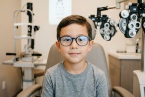 Child wearing glasses, seated in an optometrist's exam chair with a phoropter and eye chart nearby.